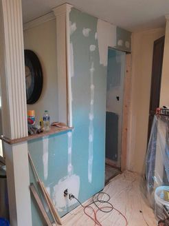 Interior wall under construction; blue-green drywall with unfinished seams, electrical outlet, and doorway.