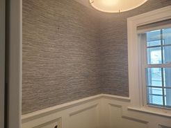A room corner with grey textured wallpaper above white wainscoting and a window with a closed blind.