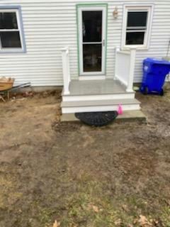 Small concrete porch with steps leading to a door. White railing and trim, blue trash bin to the side.
