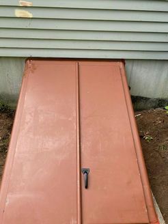 Brown metal cellar doors leading into a basement, set against green siding and a concrete foundation.