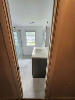 Bathroom viewed through a doorway: gray vanity with sink, window, and shower behind glass door.