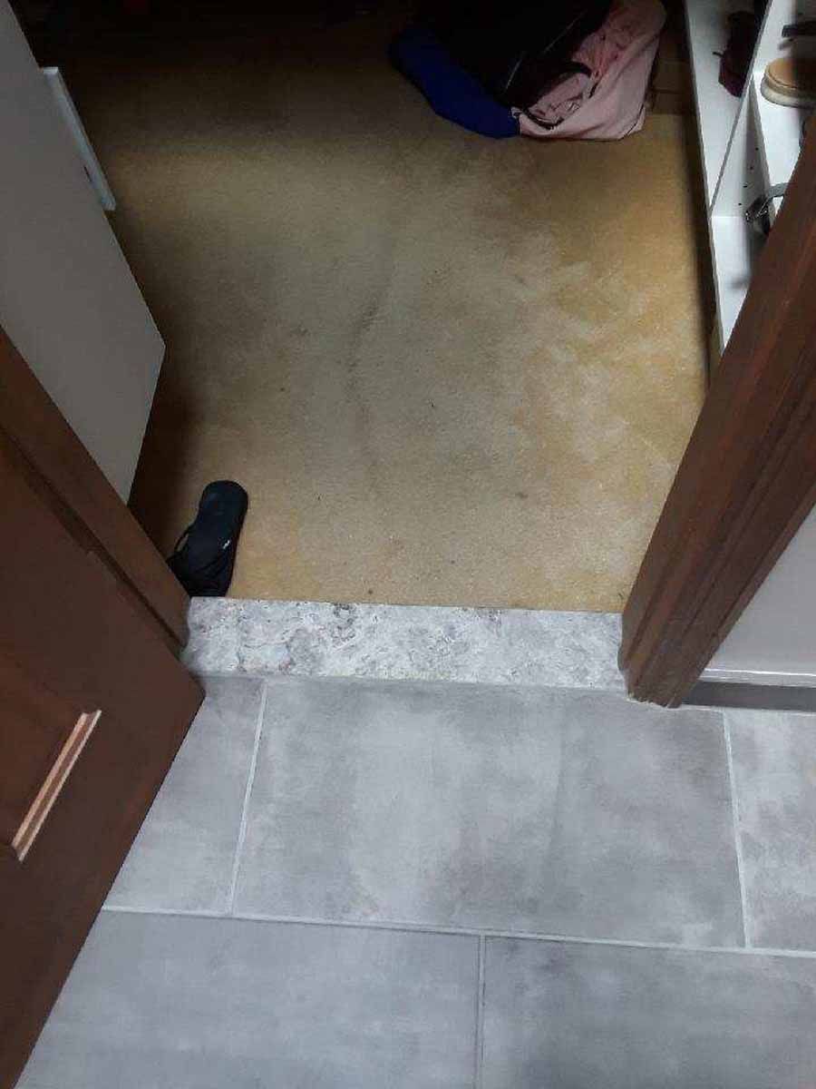 Doorway with gray tile floor leading to a room with worn, beige carpet and shoes.