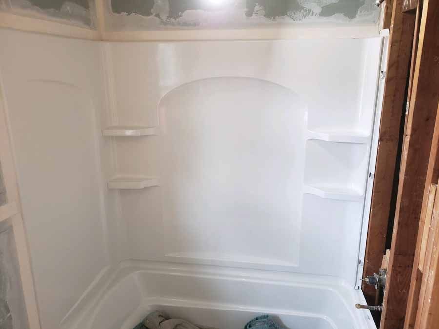 White shower unit with built-in shelves, partially installed, framed by exposed wooden studs.