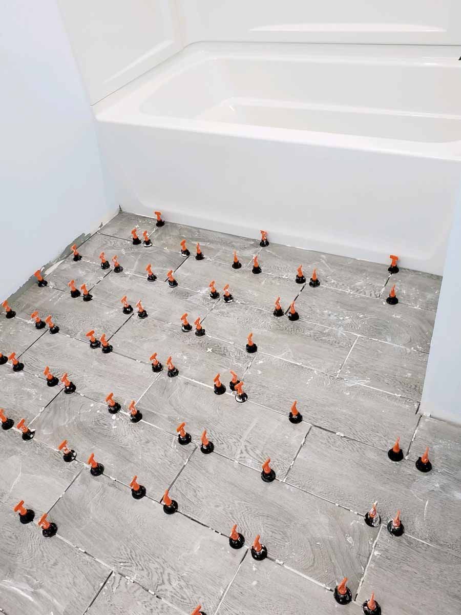 Bathroom floor with gray tiles being installed using orange leveling clips. A white bathtub is in the background.