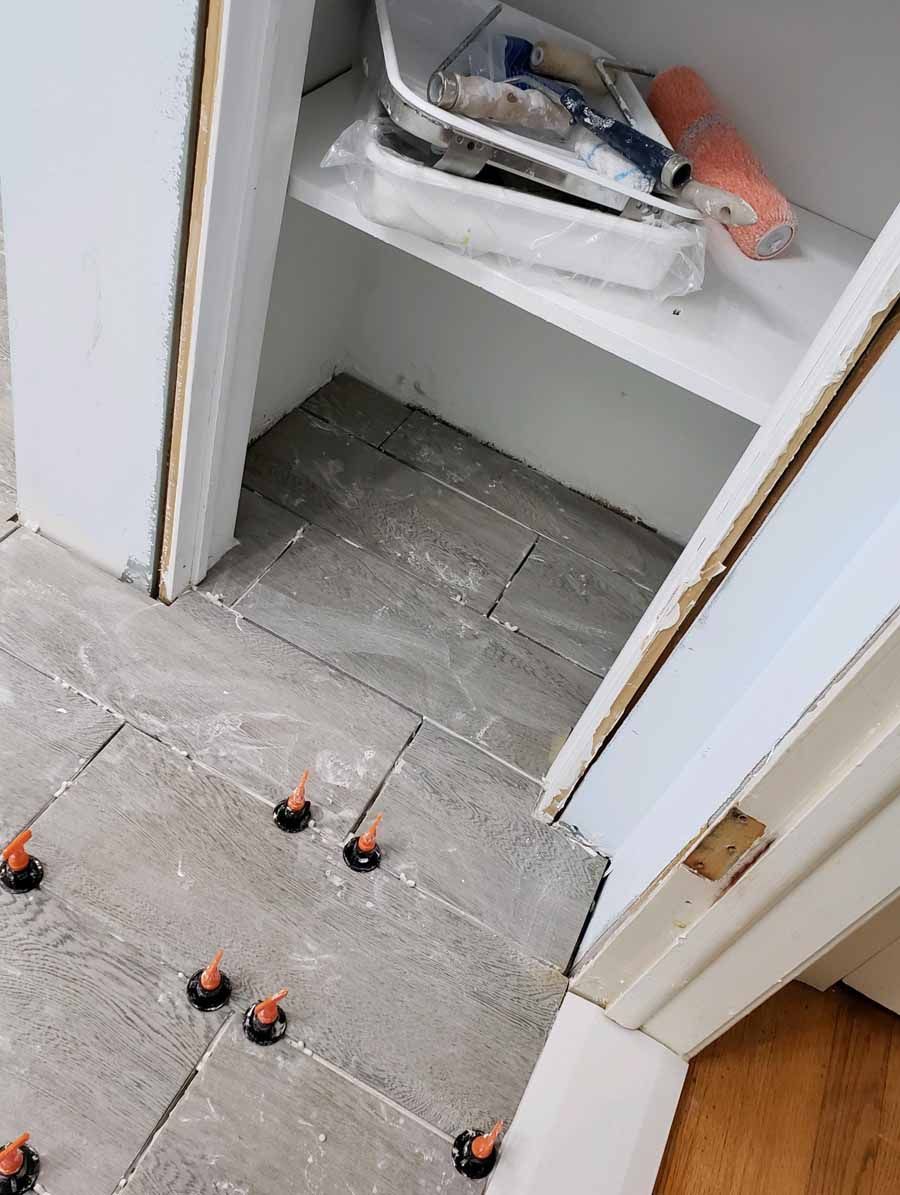 Tiled floor in progress with leveling clips. Paint supplies on a shelf above.