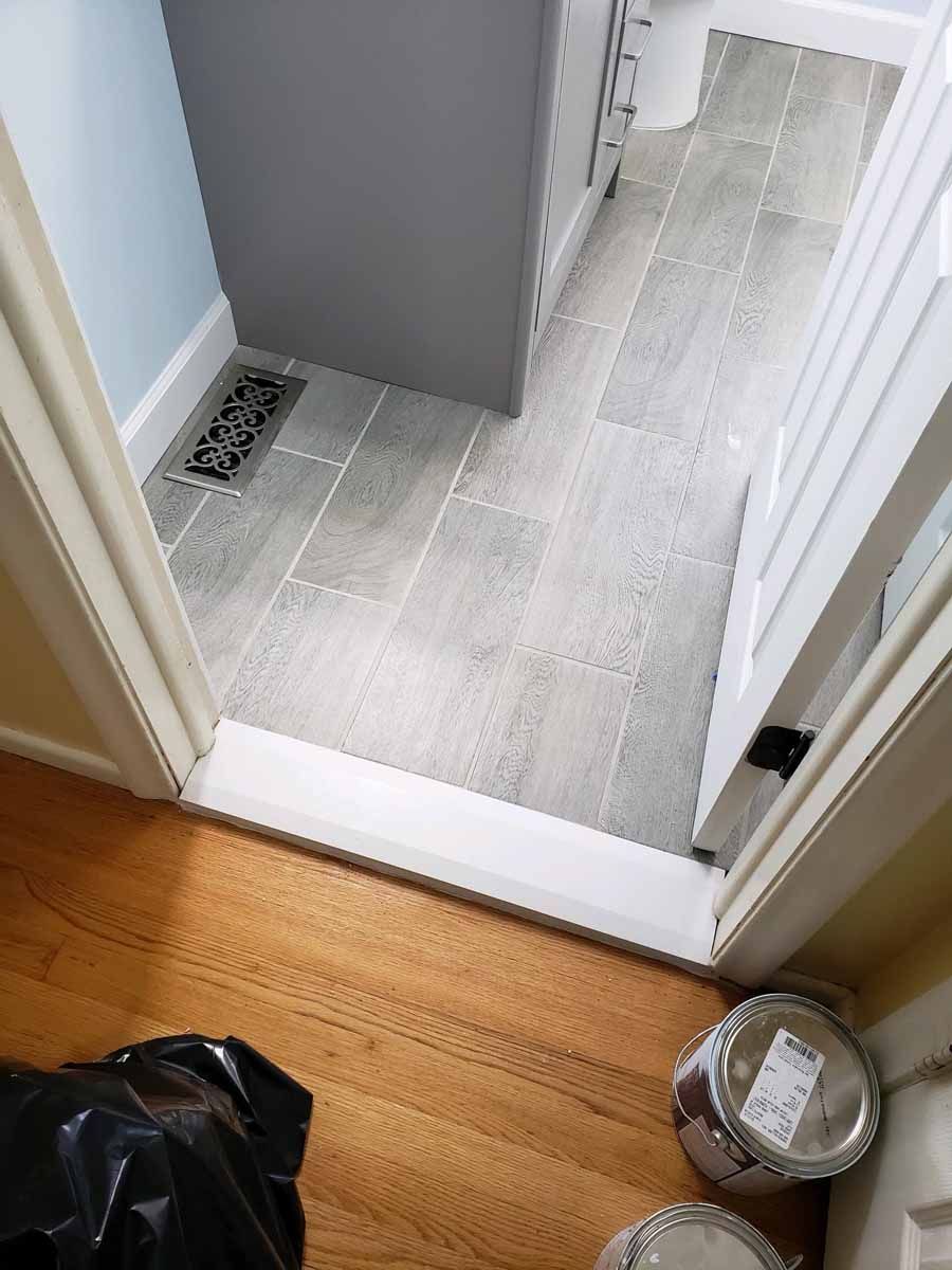 View of a bathroom entrance with wood flooring and gray tile. A black trash bag and paint cans are visible.