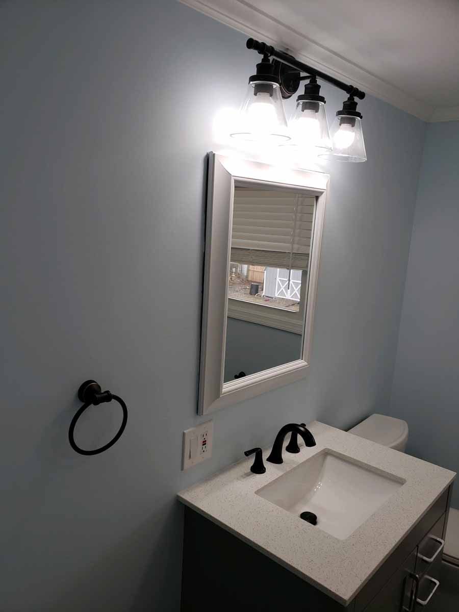Bathroom with light blue walls, vanity, mirror, and toilet. Black fixtures and a three-light vanity light.
