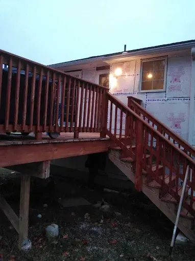 Wooden deck with stairs attached to a house with exposed insulation. Dusk setting.