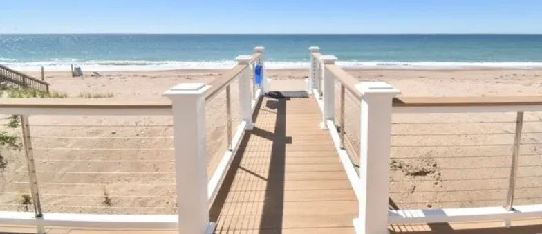 Wooden walkway with white railings leads to a sandy beach and ocean under a blue sky.