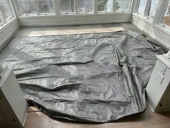 A large gray tarp covers the floor of a sunroom with many windows.