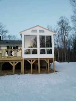 White sunroom with large windows, built on wooden supports attached to a deck, with snow on the ground.