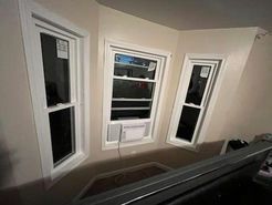 Three white-framed windows on beige walls. Air conditioner in middle window. Dark floor.