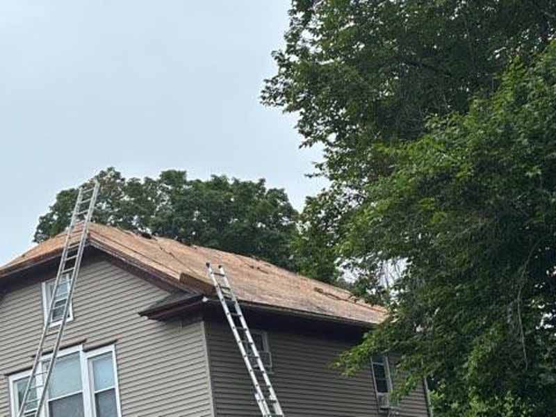 House with partially exposed roof, ladders, and surrounding trees. Overcast sky.