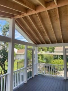 Covered porch with white railings, columns, and exposed wooden rafters, overlooking a yard with trees.