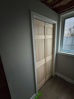 Wooden bi-fold closet doors, white trim, grey walls, with a window and a glimpse of the outdoors.