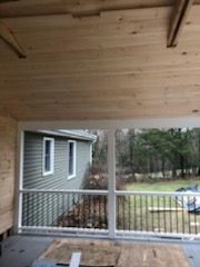 Screened porch interior with wood ceiling, overlooking backyard with house and trees.