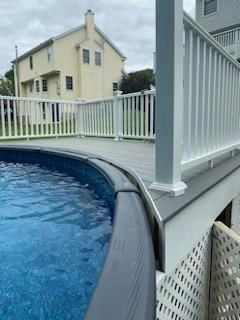 Above ground pool with blue water next to a gray deck with white railing. A light yellow house is in the background.