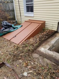 Brown cellar door with slanted metal doors, next to a concrete well, against a yellow house.