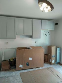 Kitchen renovation in progress: gray upper cabinets, cardboard boxes on the floor, unfinished walls, and a ceiling light.