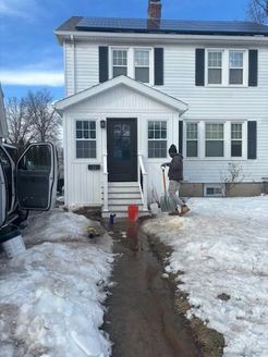A person digging a trench in front of a white house with black shutters, snow on either side.