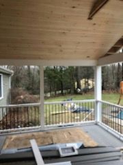 Covered porch with wood ceiling and railings, overlooking a backyard with trees.