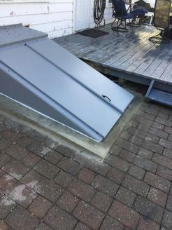 Gray cellar door on brick patio, leading to a wooden deck with a chair and hose.