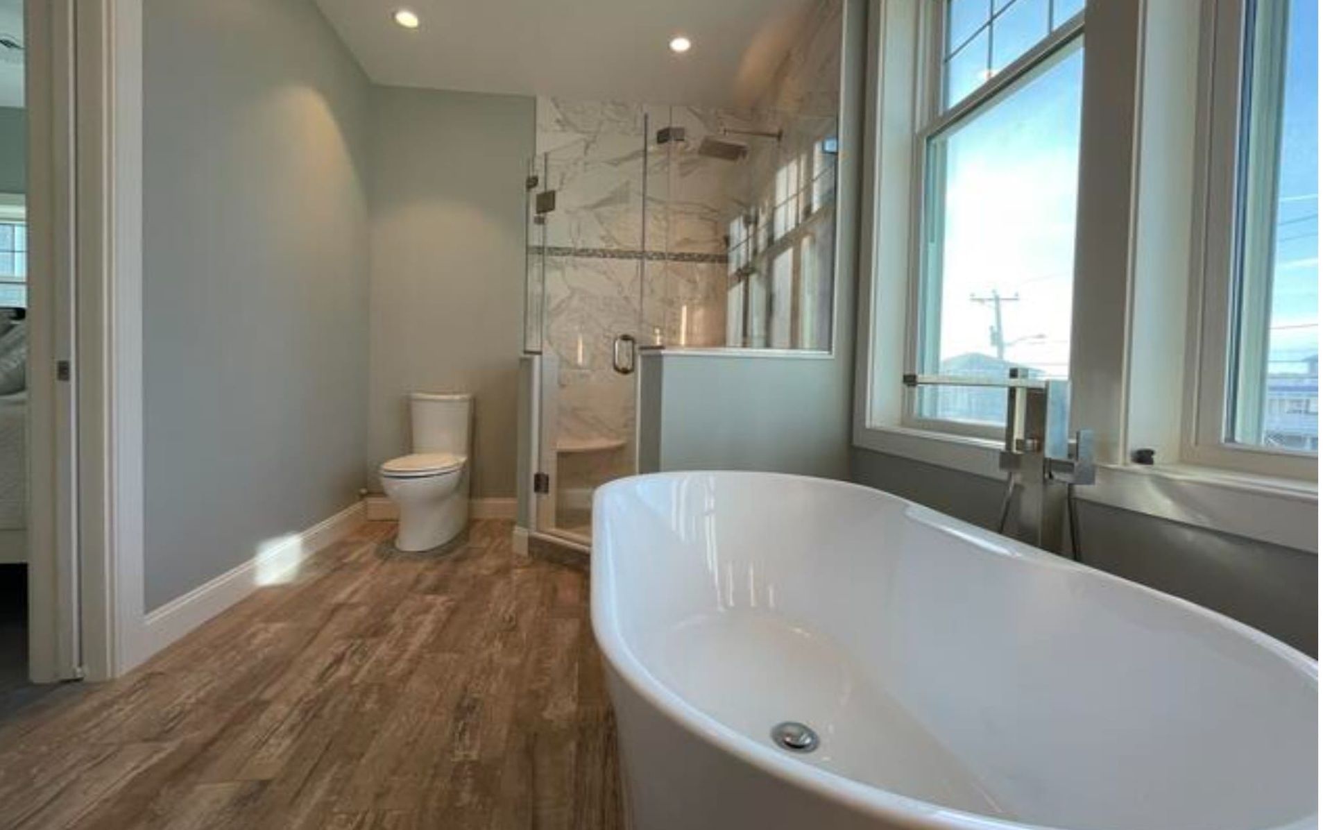Bathroom with a white tub, toilet, and shower. Wooden-look floor, and large window.