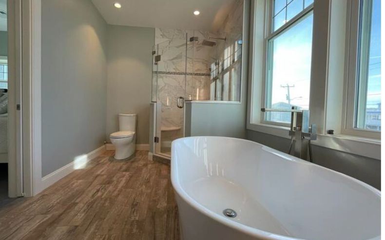 Bathroom with a white tub, toilet, and shower. Wooden-look floor, and large window.