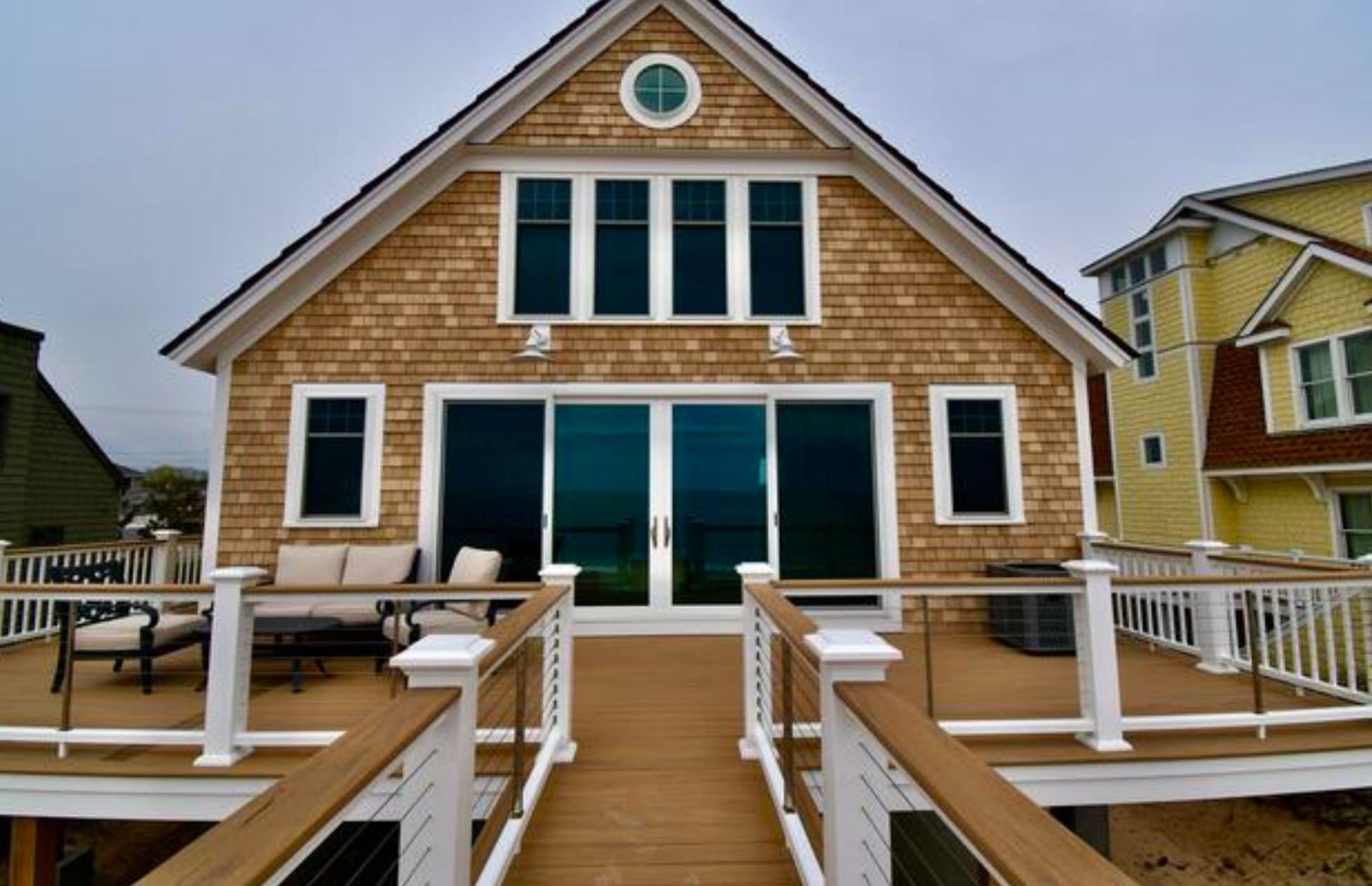 Beach house with brown shingles, deck, and large glass doors, facing the ocean.