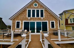 Beach house with brown shingles, deck, and large glass doors, facing the ocean.