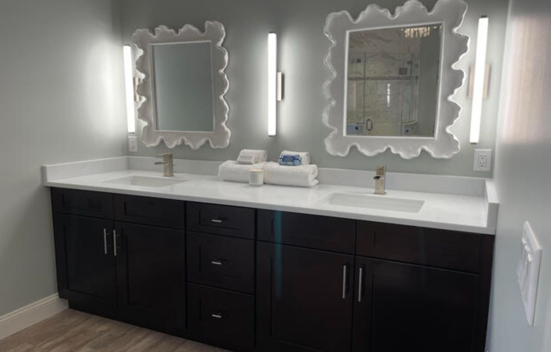 Bathroom with two sinks, dark cabinets, white countertop, wavy-edged mirrors, and vertical lights.