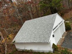 White garage with gray shingled roof, two doors, surrounded by autumn trees and foliage.