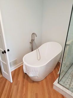 Freestanding white bathtub in a corner of a bathroom with wooden floors and a glass shower.
