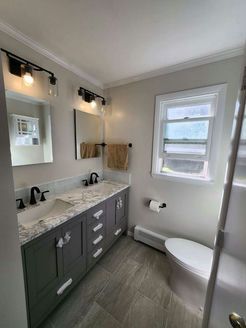 Bathroom with gray cabinets, double sinks, and a toilet. Light fixtures and a small window are visible.