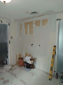 Room under renovation, walls partially stripped with exposed drywall, floor debris, tools present.