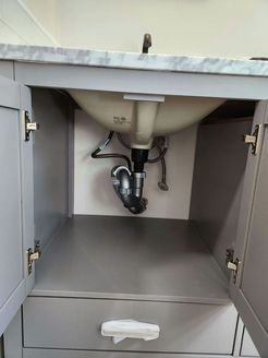 Under-sink cabinet with open doors, showing plumbing and a white basin. Gray cabinet, marble countertop.