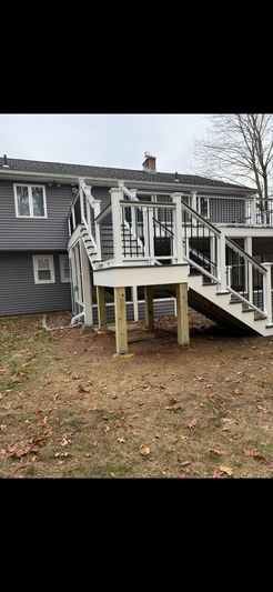 Backyard view of a gray house with a white deck, stairs, and exposed wooden supports, in a yard with dry leaves.