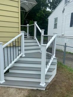 Gray deck stairs with white railings and black spindles, leading to a yellow house with a white awning.