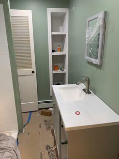 Bathroom with new white vanity, built-in shelving, and pale green walls. A mirror is wrapped in plastic.