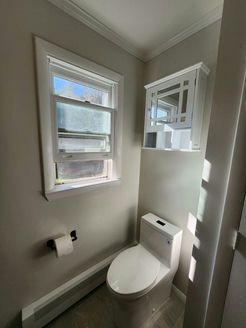 A small bathroom with a toilet, window, cabinet, and toilet paper roll. Gray walls, white trim.
