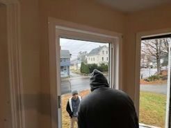 Two people by a window, one in a hood, looking outside at a street with houses and autumn foliage.