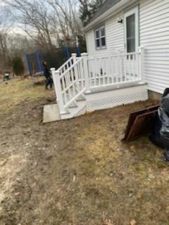 White deck with railing next to a house with a grassy yard. A person is near a trampoline.