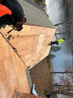 Roofers working on a building with a water view. One uses a hammer. Another wears a neon vest.