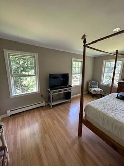 Bedroom with hardwood floors, two windows, a four-poster bed, and a TV on a stand. Walls are a neutral color.
