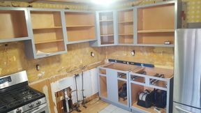 Kitchen cabinets being installed; gray frames, natural wood interiors, stainless steel appliances, construction setting.