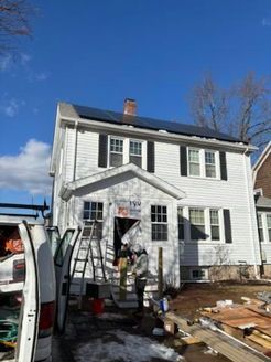 Solar panels on a white house roof. Construction workers on porch. Van parked nearby. Blue sky.