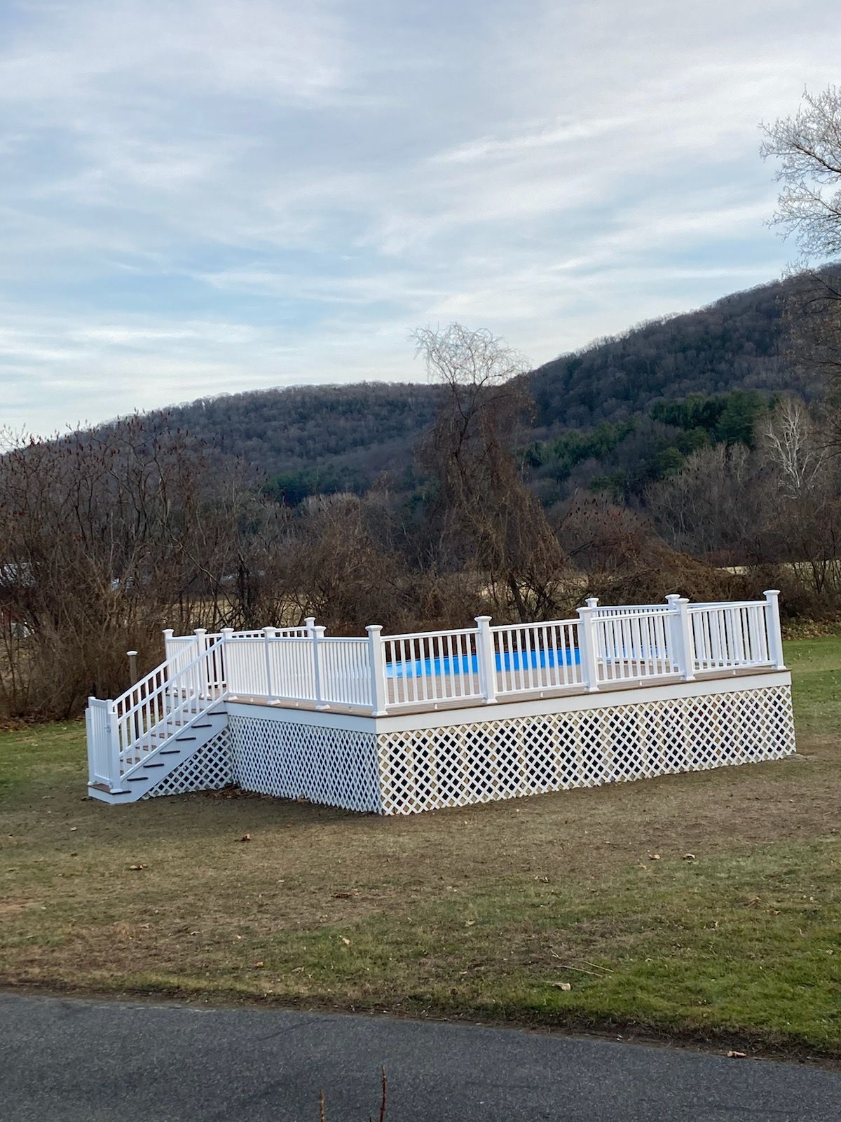 White-fenced deck with blue pool, on a grassy lawn with hill backdrop, under a cloudy sky.