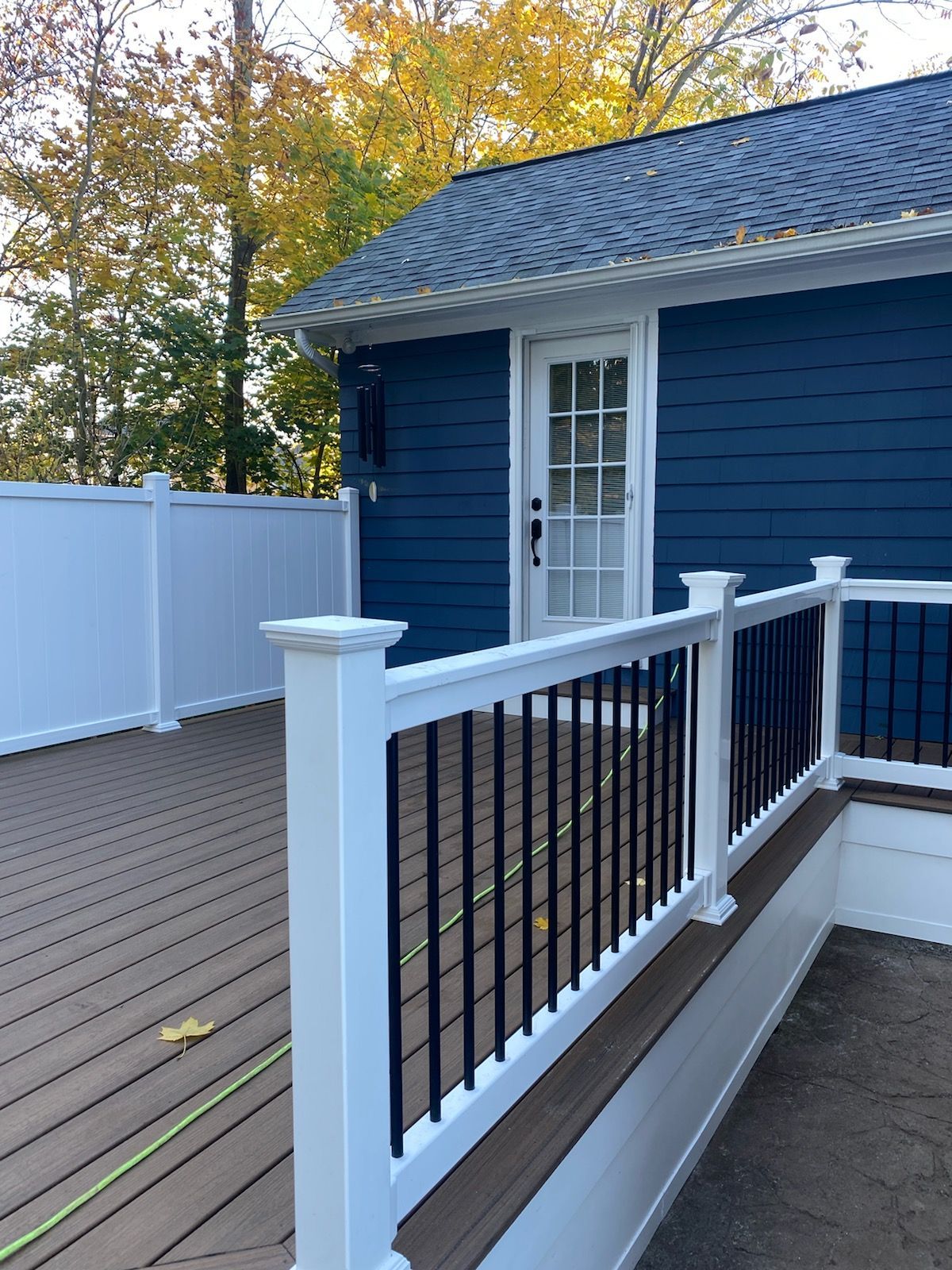 Deck with white and black railing, adjacent to a dark blue house with white door; autumn foliage in the background.