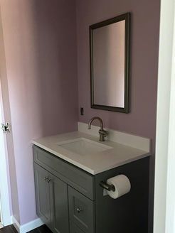 Gray bathroom vanity with white countertop, chrome faucet, and framed mirror on a lilac-painted wall.