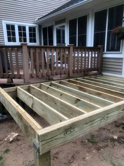Wooden deck frame under construction next to a house with a completed deck and windows.
